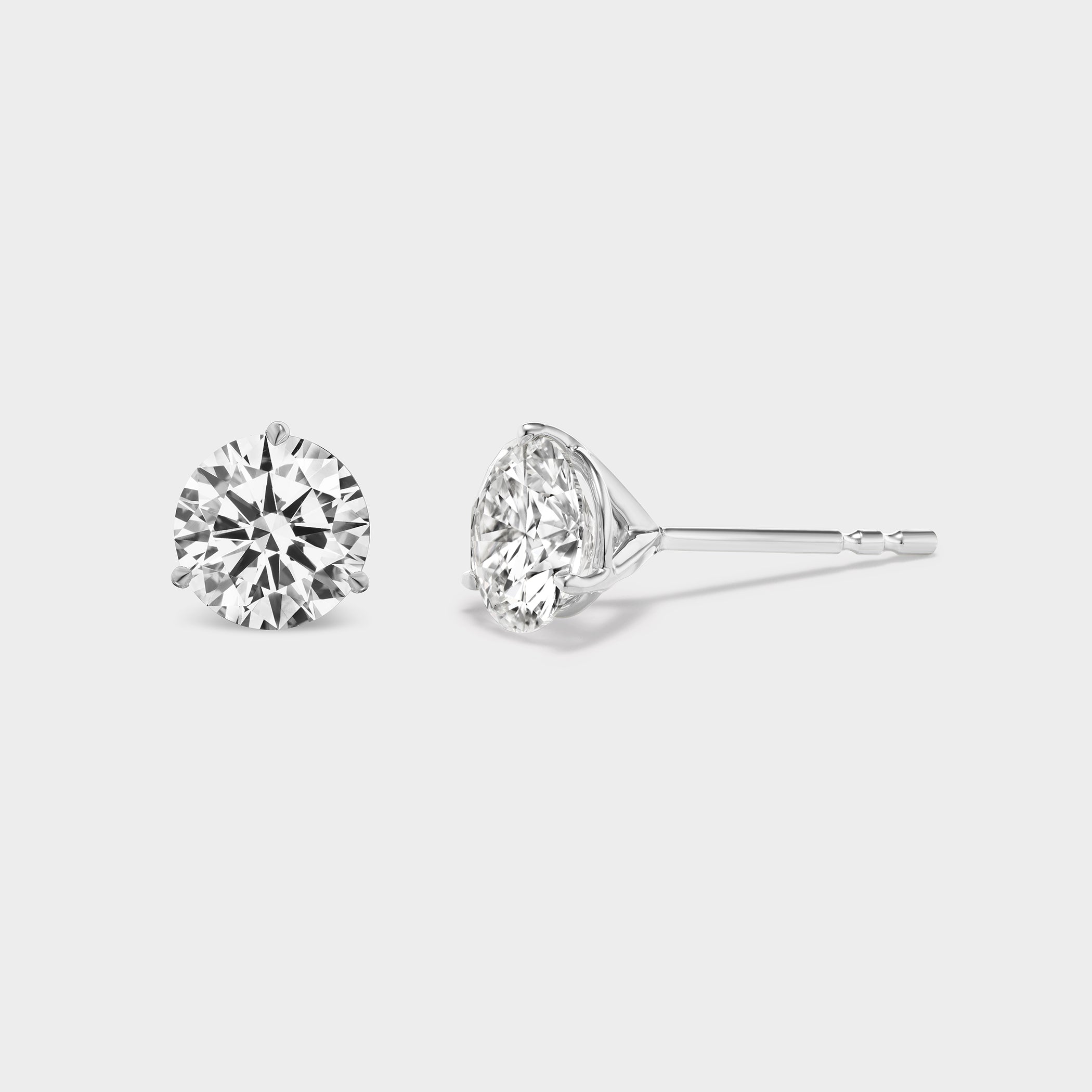 3 Prong Martini Diamond Studs. Close-up side view of a pair of round diamond earrings on a neutral background. Everyday sized lab diamond stud with modern martini setting in 14K white gold perfect for building an ear stack.