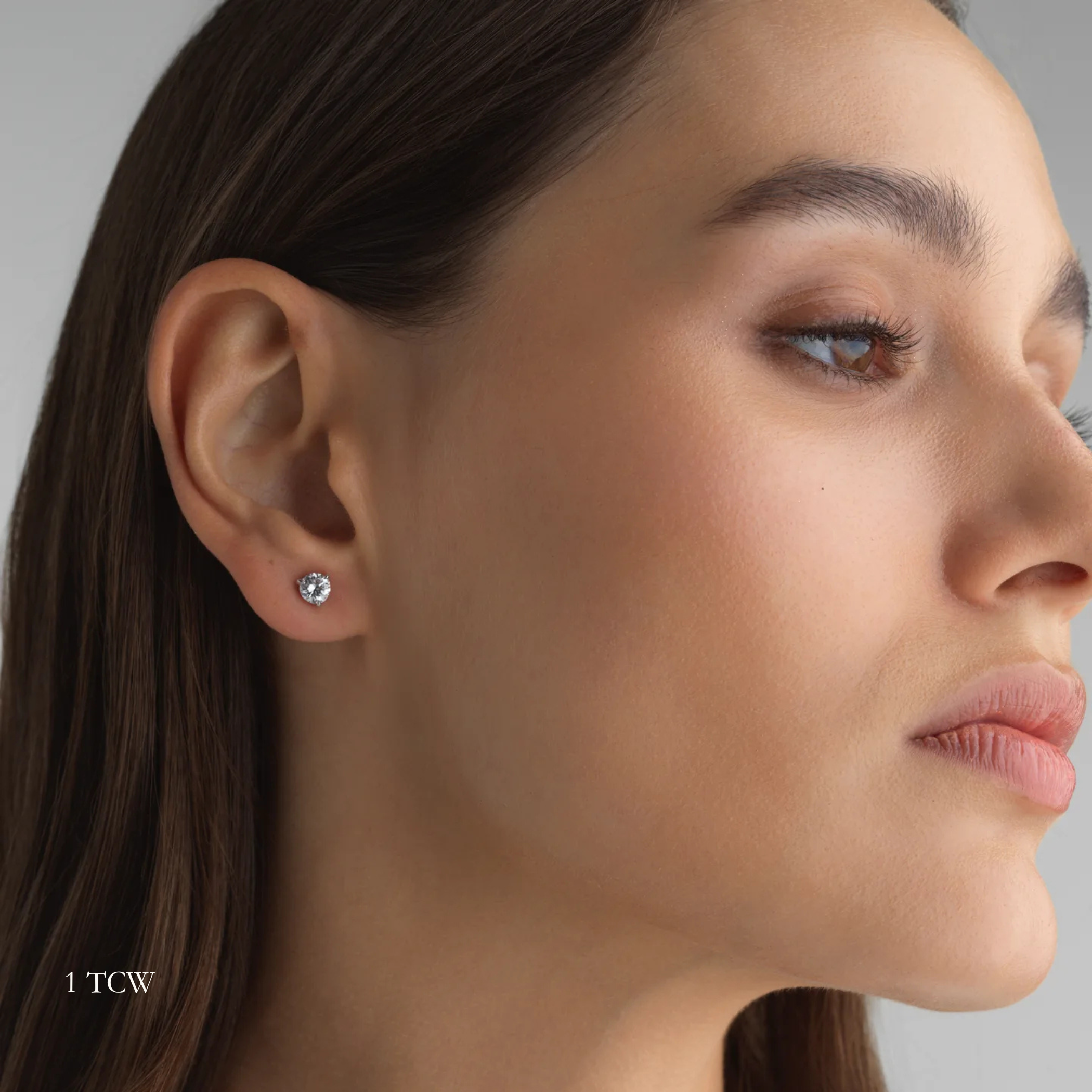 Close-up of a woman wearing a 1 total carat round diamond earring on a neutral background. Everyday sized lab diamond stud with modern setting in 14K gold for work or school.