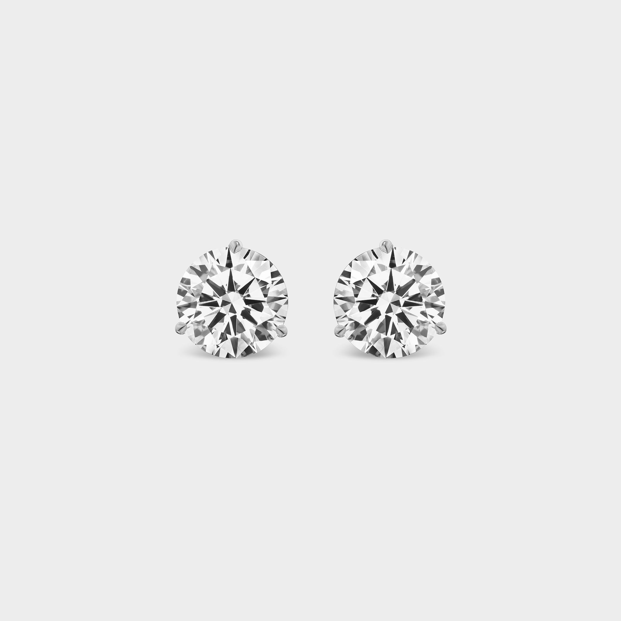 3 Prong Martini Diamond Studs. Close-up of a pair of round diamond earrings on a neutral background. Everyday sized lab diamond stud with modern martini setting in 14K gold perfect for building an ear stack.