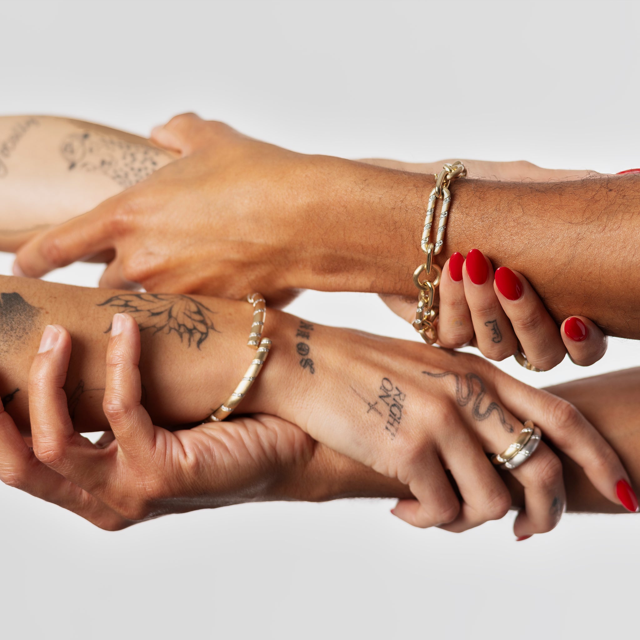 Four diverse hands interlocked, showcasing a gold chain bracelet, a dome ring, and various tattoos.