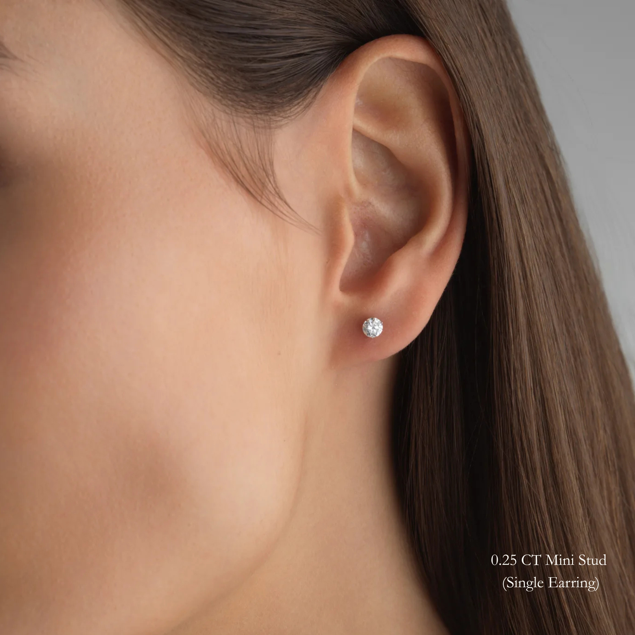 3 Prong Martini Diamond Studs. Close-up view of a woman wearing a 0.25 CT round diamond earring in 14k gold on a neutral background. Everyday sized lab diamond stud with modern setting in 14K gold perfect for building an ear stack