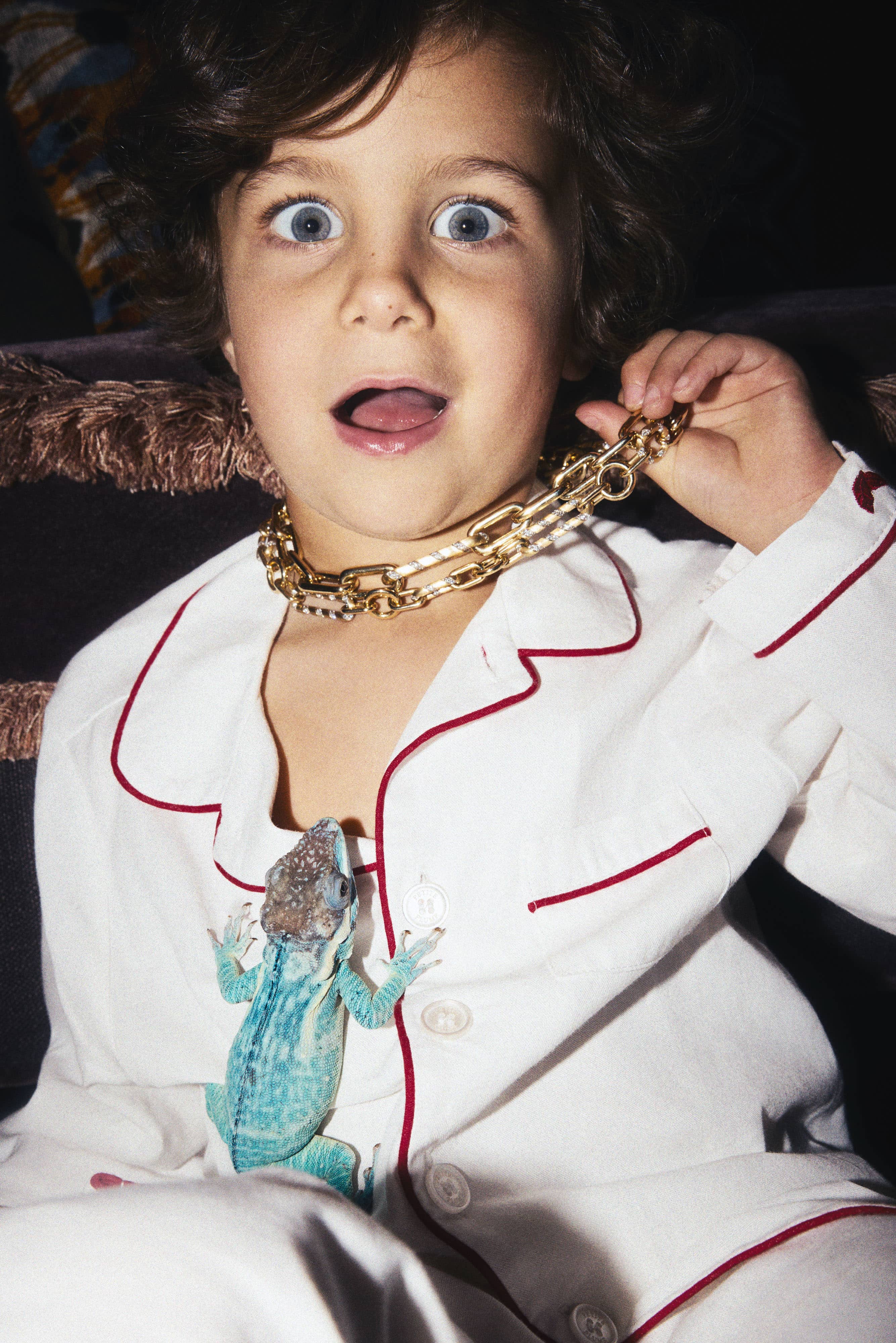Child with wide eyes wearing a gold mixed link necklace and white pajamas, a blue lizard on their chest.