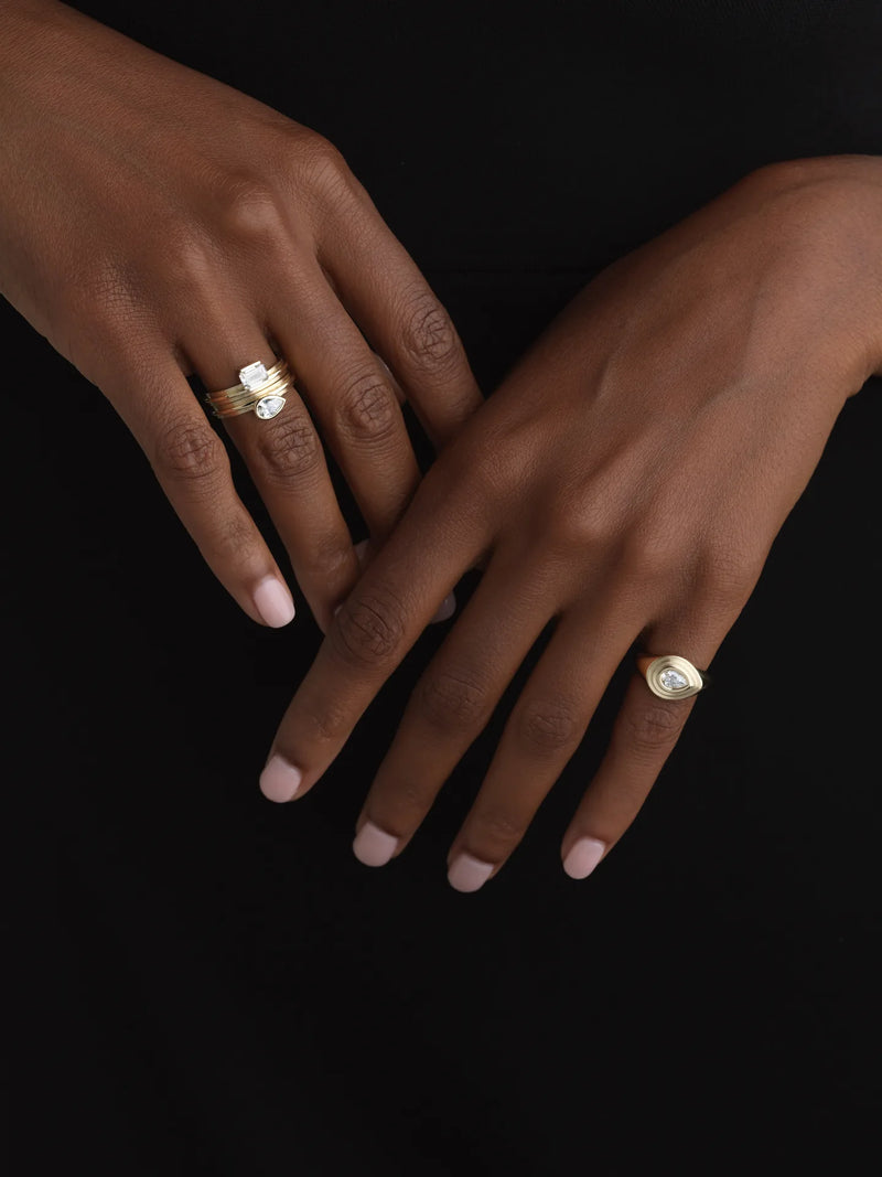 Two hands with light pink manicures wear 14K gold rings. One hand wears a pear-cut lab-grown diamond ring, the other a modern oval ring.