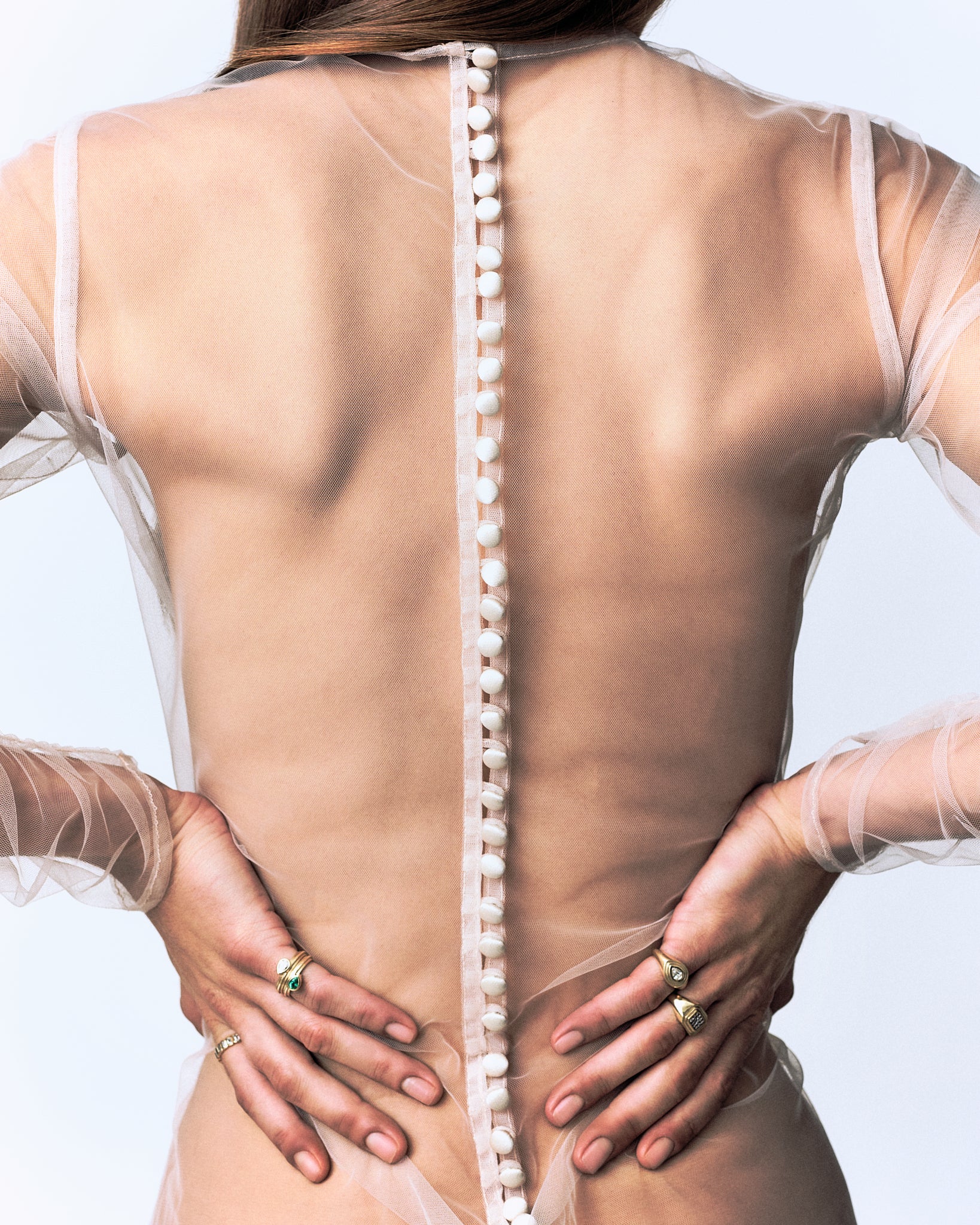 Woman's back in sheer dress with pearl buttons, hands on hips, wearing gold rings with pear and emerald stones.